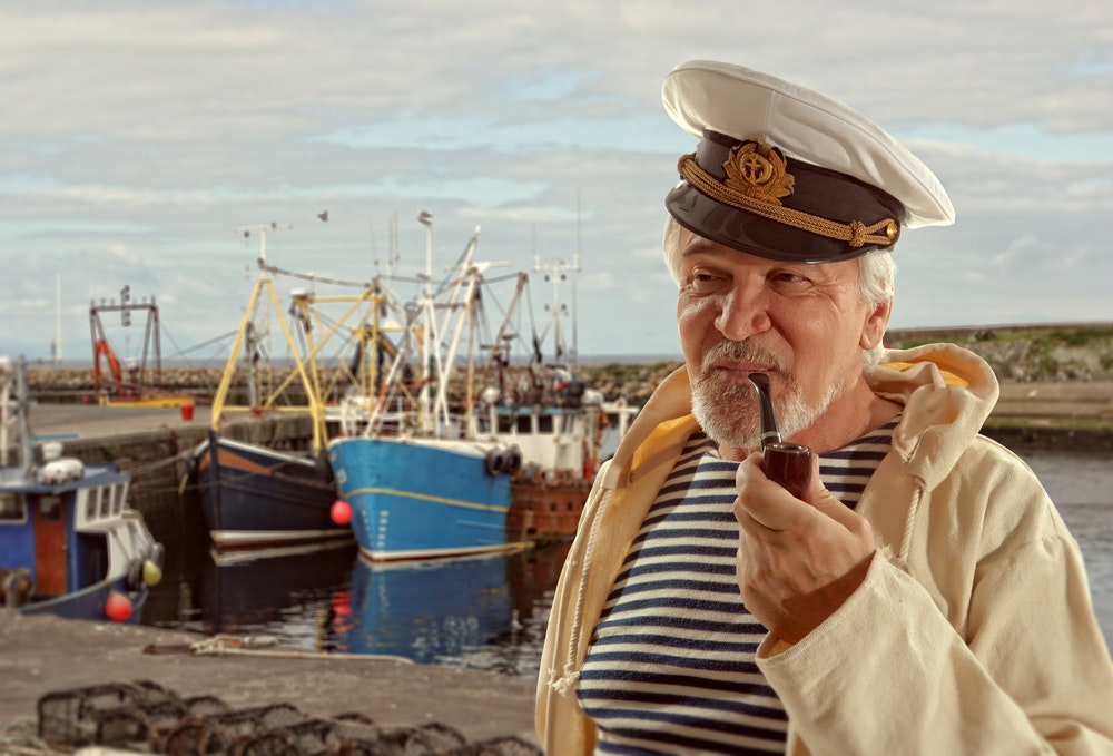 En eldre, gråhåret mann med kapteinlue og pipe i munnen.