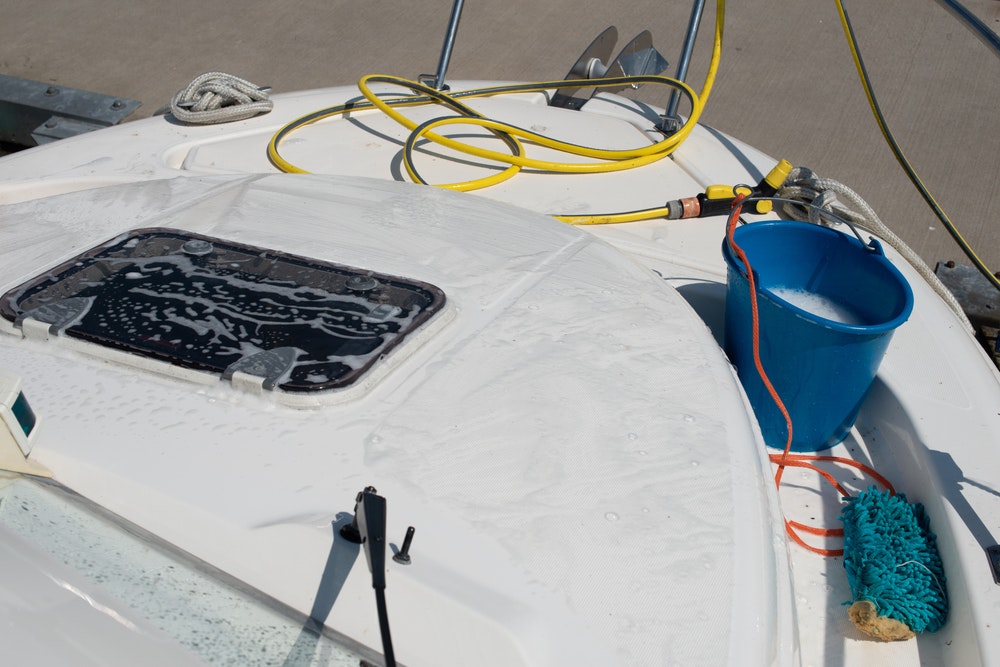 Laivų valymo įranga. Ant jachtos guli plastikinis kibiras, žarna su vandens purkštuvu, šluota.