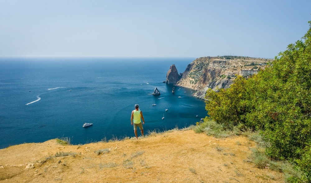 海辺の崖をハイキングする男性、湾に浮かぶボートの眺め。
