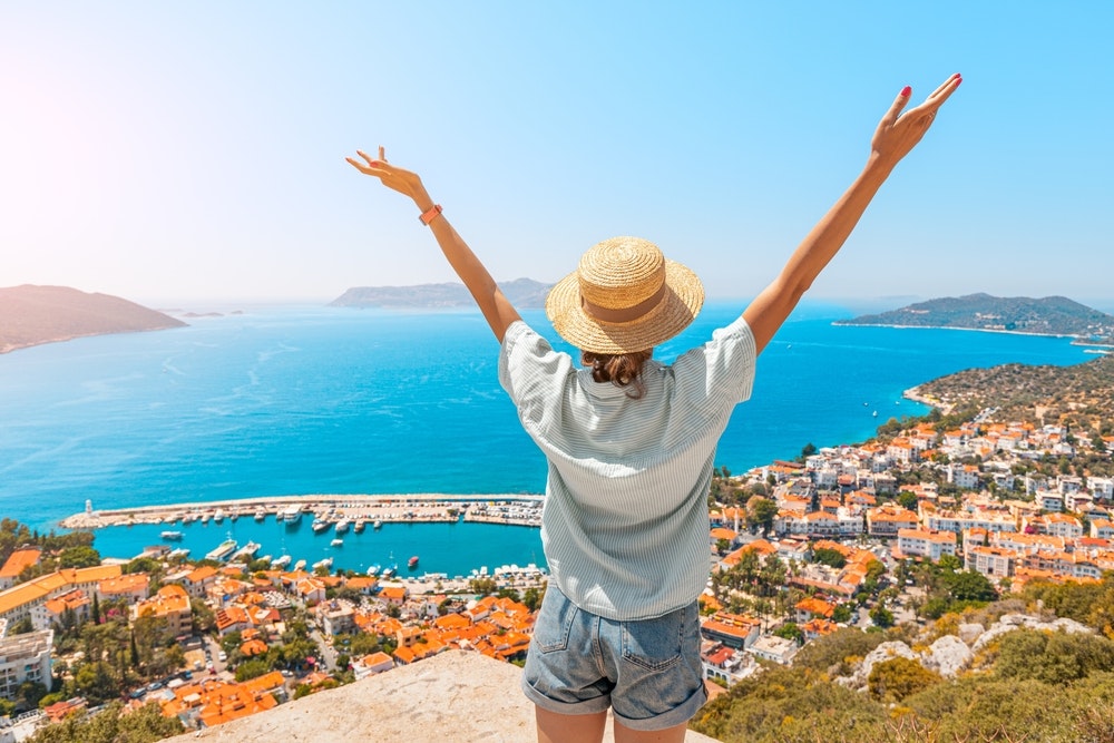 Laimīga sieviete ar atplestām rokām stāv uz skatu punkta, baudot Kasas kūrorta panorāmu pie Vidusjūras Turcijā.