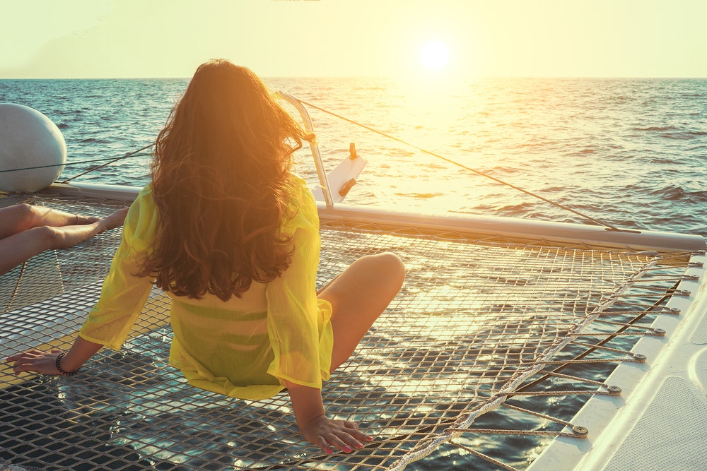Een vrouw zit op een net op een catamaran en kijkt met een naar de zonsondergang.