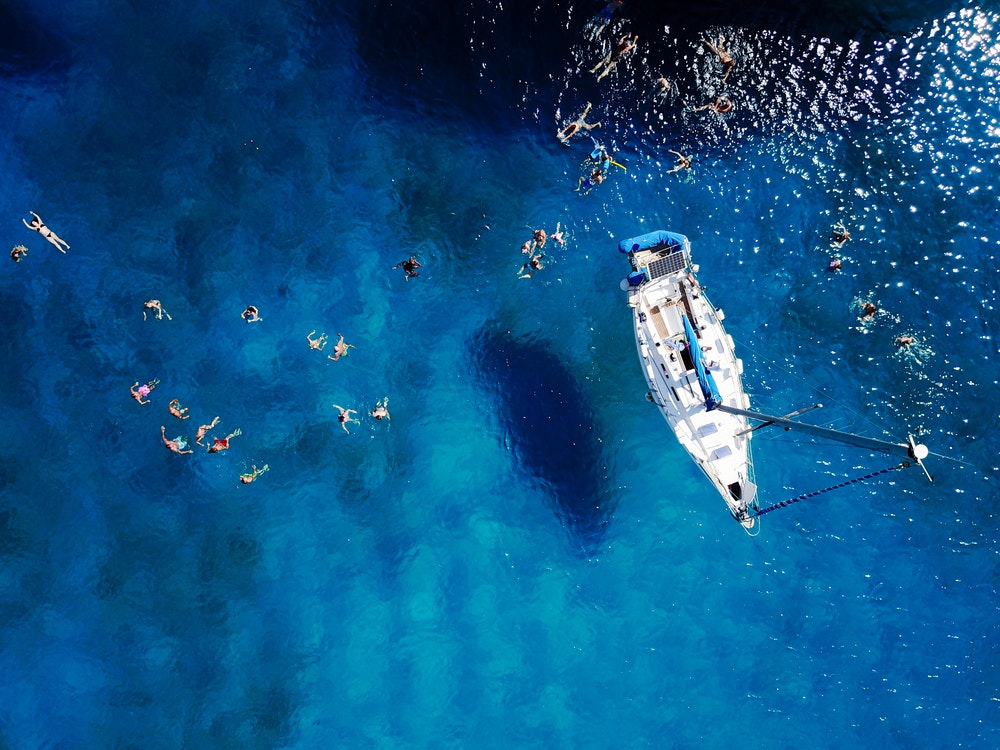 Skats no gaisa uz skaistu zilu lagūnu karstā vasaras dienā ar buru laivu. Skats no augšas uz cilvēkiem, kas peld ap laivu.
