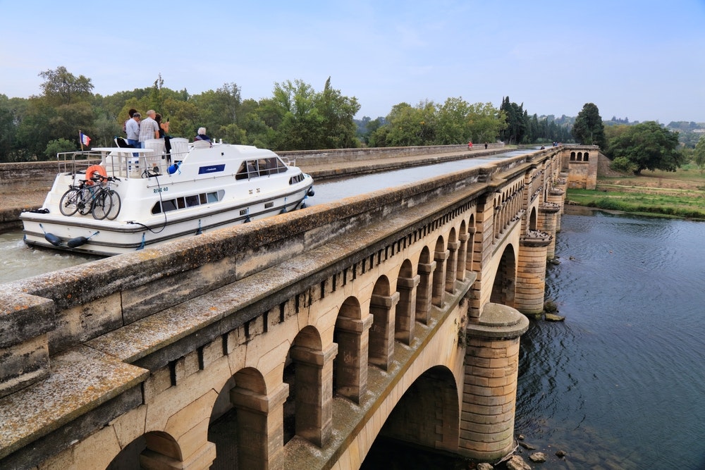 Лодка пересекает мост через реку Орб на историческом Канале дю Миди во Франции. Канал дю Миди является объектом Всемирного наследия ЮНЕСКО.
