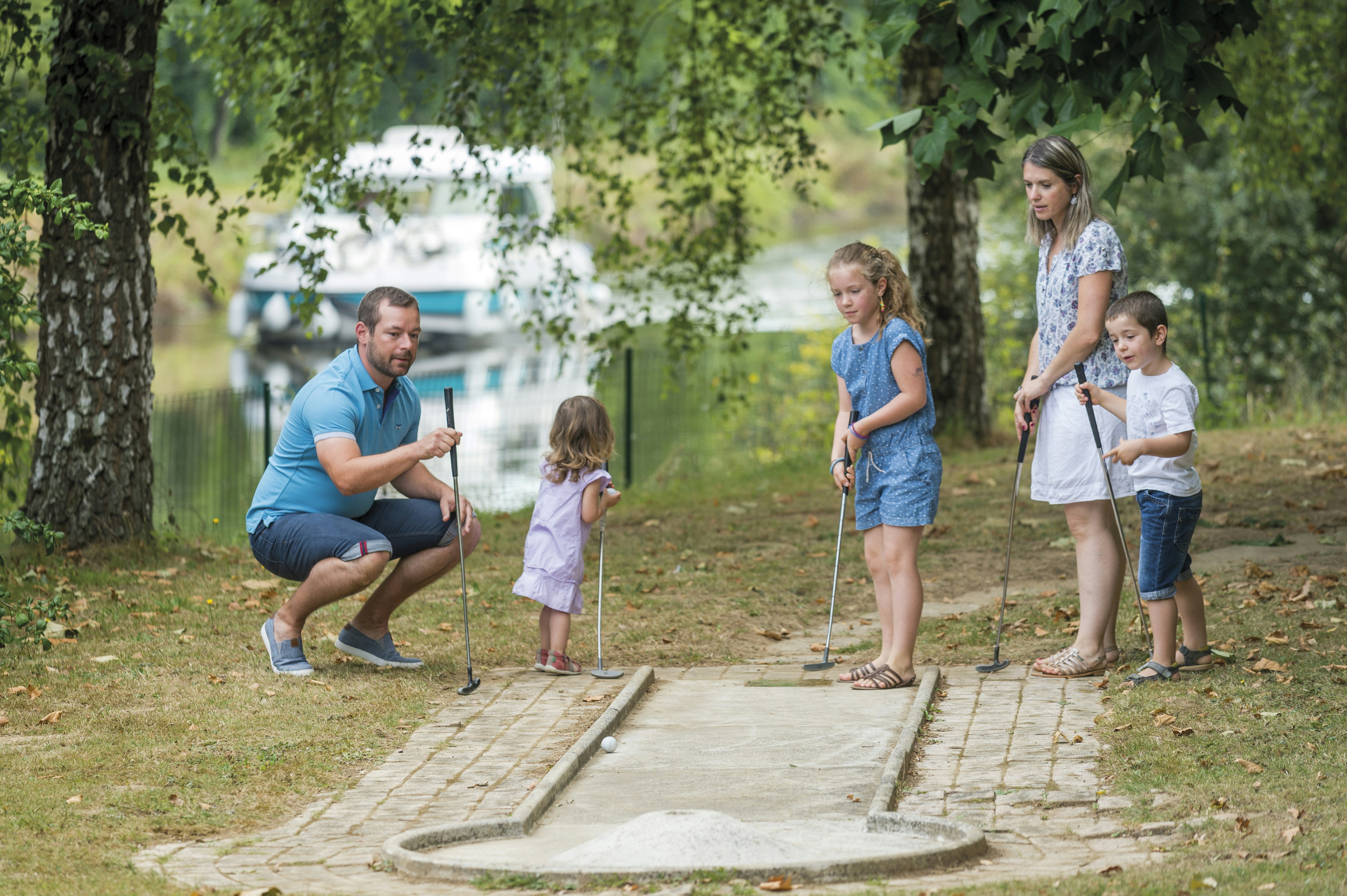 Družina igra mini golf z vodnim kanalom in hišnim čolnom v ozadju