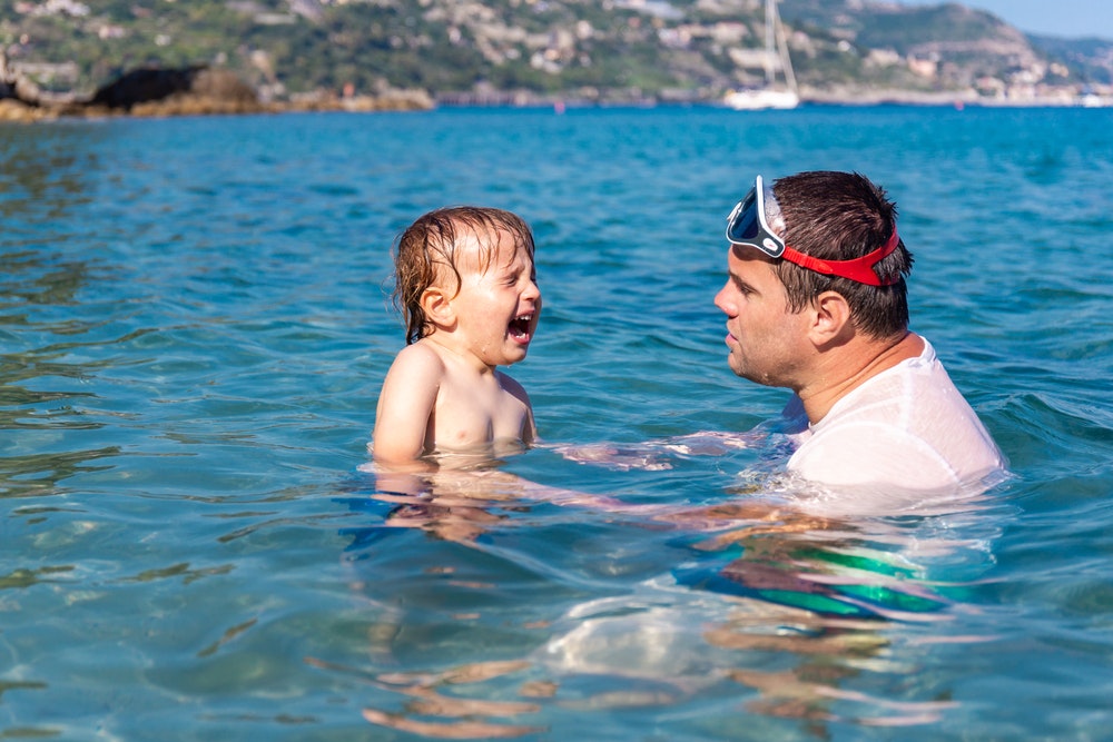 Ett litet barn gråter och är rädd för vattnet i havet. Far och son simmar tillsammans i havet under sommarlovet.