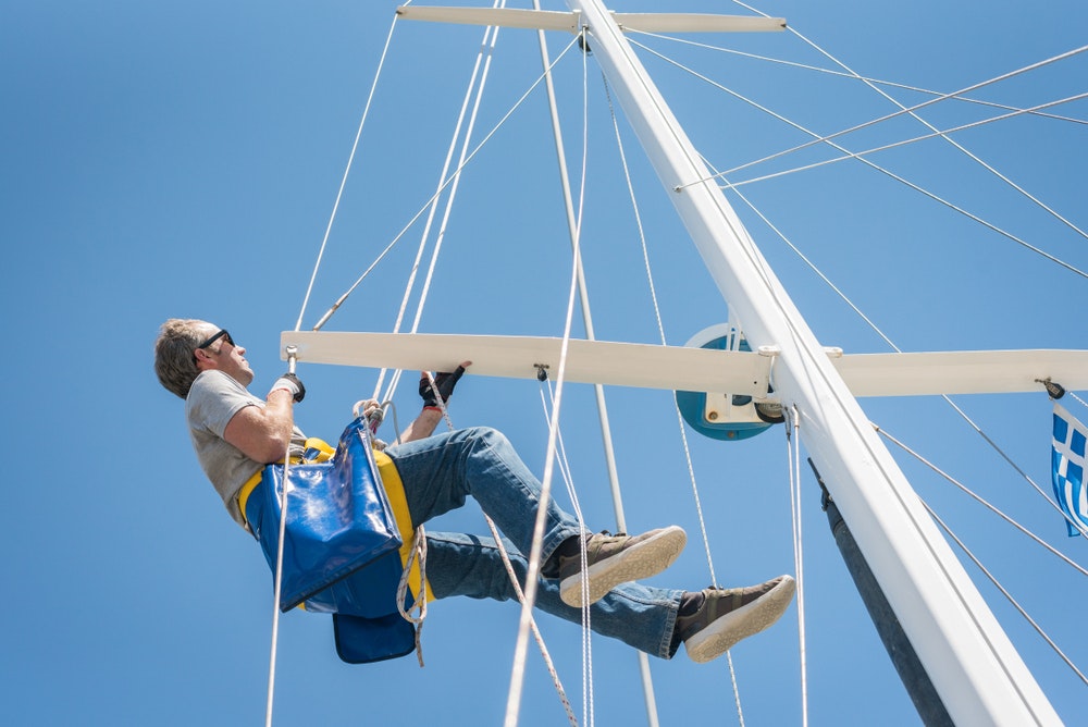 Caucasian Male working up the mast of a sailing yacht, with rope and bosun's chair on a sunny day with blue sky