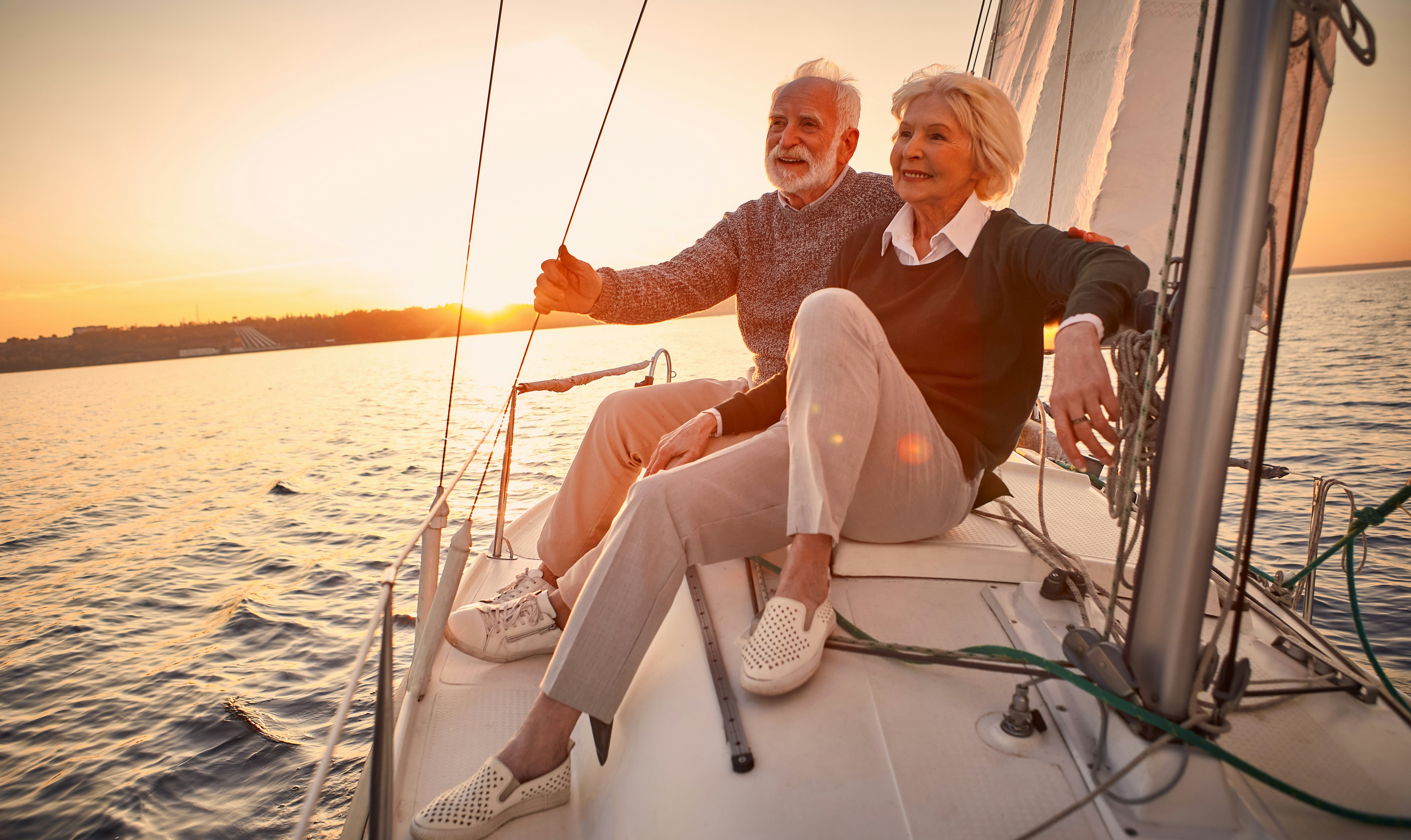 A beautiful and happy senior couple in love sitting on the side of a sailboat or yacht deck floating in the sea at sunset and enjoying the amazing view, sailing together