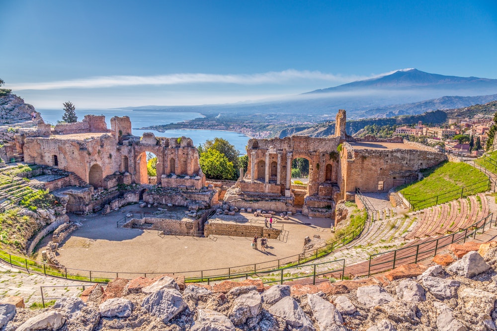 Die Ruinen eines antiken griechischen Theaters in Taormina, Sizilien, mit dem schwelenden Vulkan Ätna im Hintergrund.