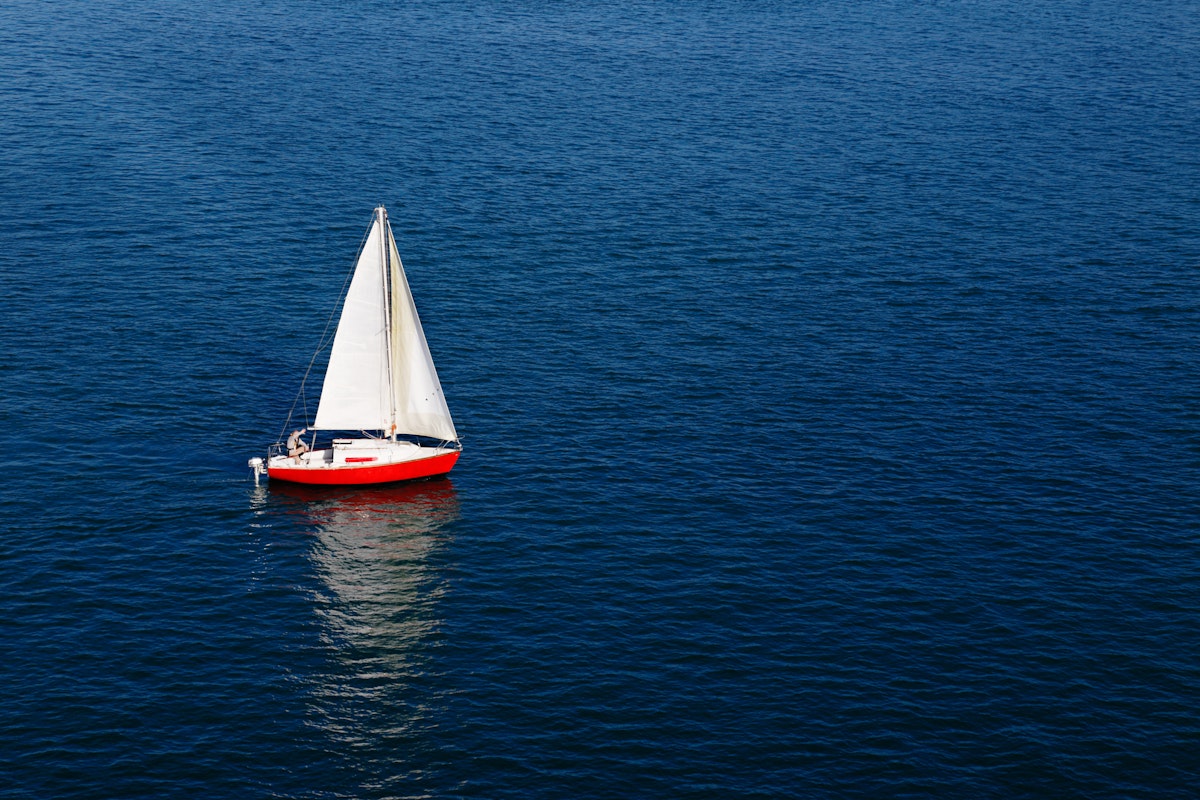 A lone white sail of a red sailboat on a calm blue sea