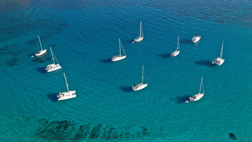 ターコイズブルーの海が広がる湾の上空から、帆船が間隔をあけて停泊している様子を眺める。