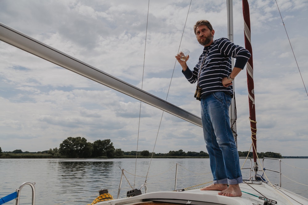 A bearded businessman in a striped sweater stands on the bow of a white yacht, sipping whisky from a glass.