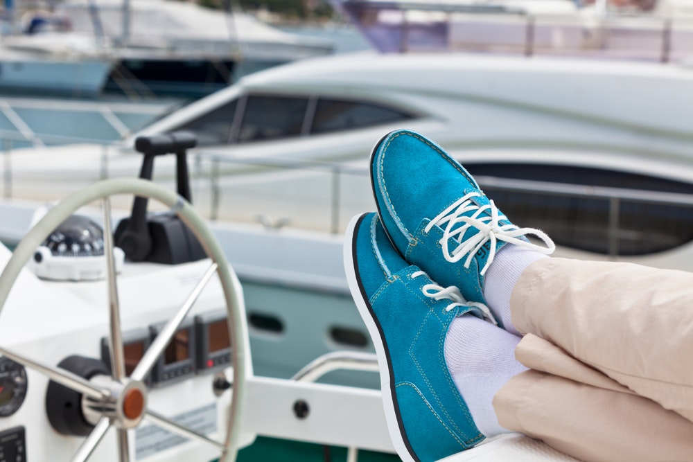 Žmogaus kojų pora su kelnėmis ir ryškiai mėlynu kombinezonu jachtos fone