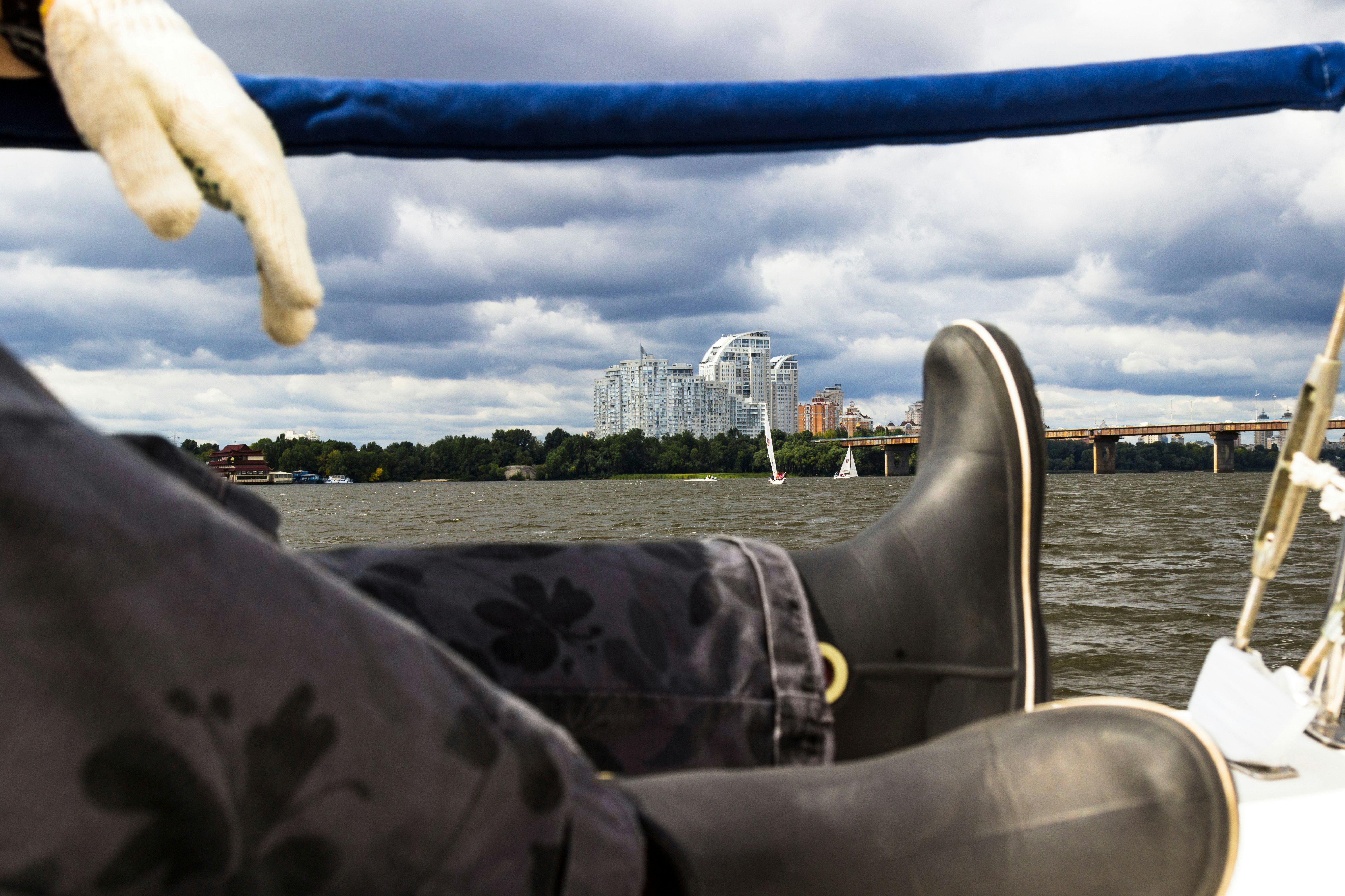 Feet in black boots aboard a yacht. Ahead is the riverbank with a cityscape. Part of the frame