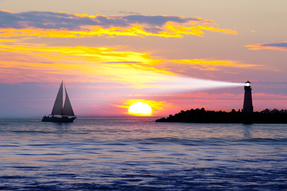 Auringonlasku merellä, purjevene ja loistava majakka.