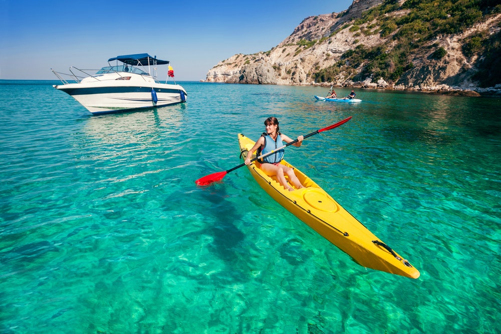 Femme en kayak sur l'eau claire d'une baie, bateau à moteur en arrière-plan. 